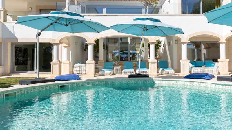 Türkisfarbener Pool mit klassischen weißen Säulen und blaugrünen Sonnenschirmen auf einer sonnigen Terrasse.