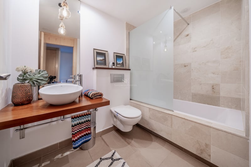 Salle de bain moderne avec plan de toilette en bois, vasque à poser, WC suspendu et baignoire carrelée en pierre.