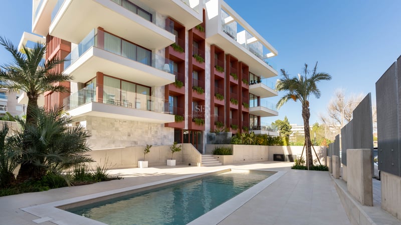 Modern apartment building facade with glass balconies and a rectangular swimming pool framed by palm trees.