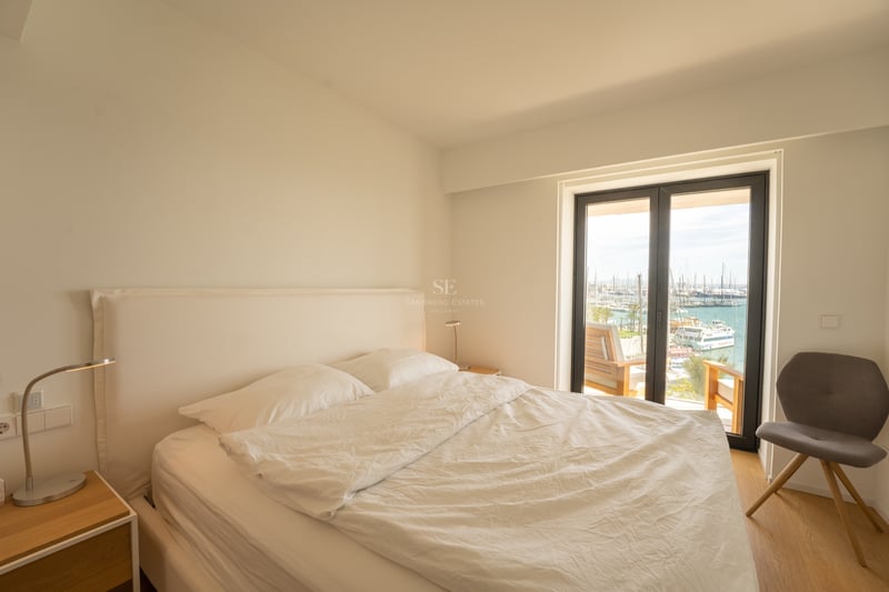 Ljust sovrum med vit säng, grå stol och stora glasdörrar mot en marina fylld med båtar.