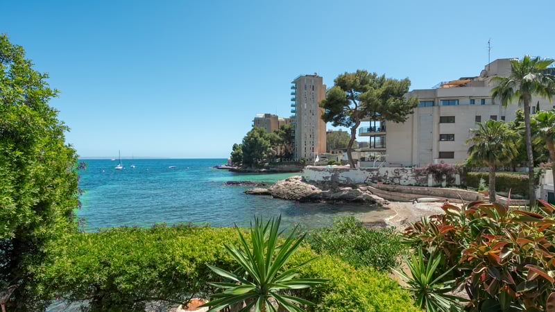Turkos vik med klippig kust och moderna byggnader inramade av medelhavsgrönska under en klarblå himmel.