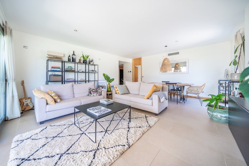 Salon spacieux avec canapés beiges, tapis en laine, étagères en métal noir et coin repas en arrière-plan.