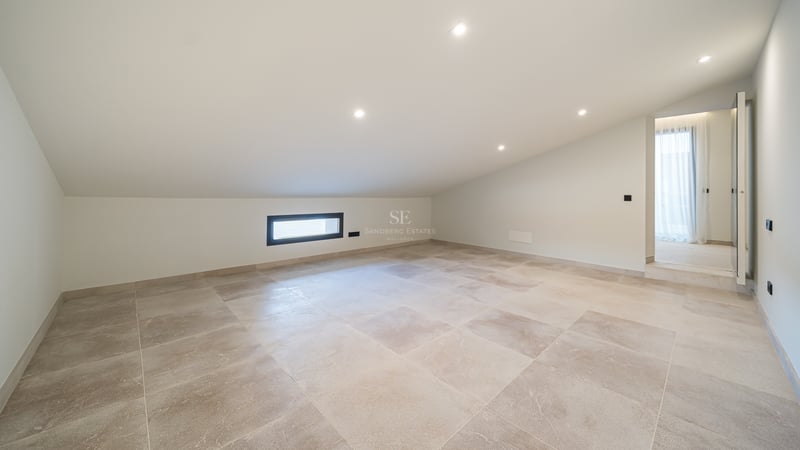 Pièce vide avec plafond incliné blanc, carrelage en pierre beige et spots encastrés.