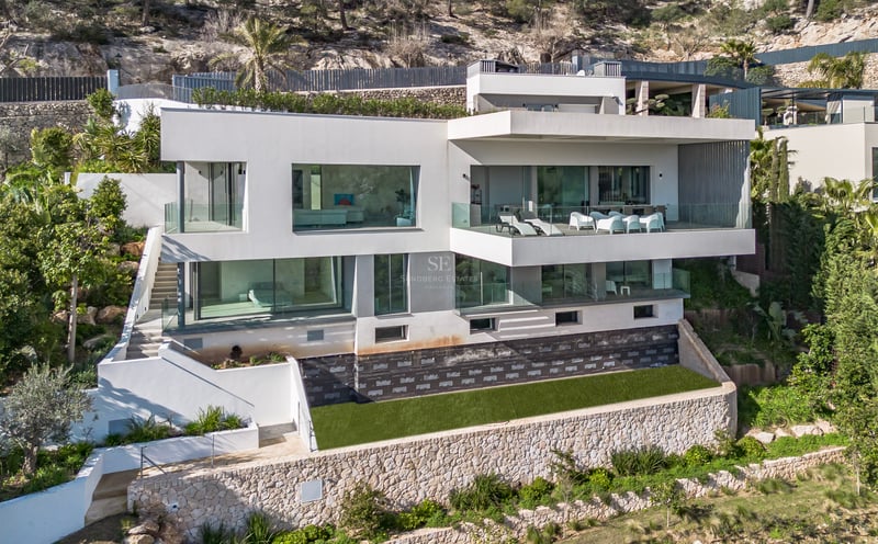 Villa blanche moderne de plusieurs étages avec de grandes baies vitrées et terrasses construite sur une colline rocheuse.