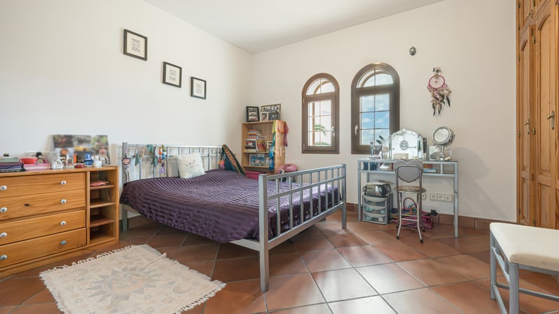 Bedroom featuring terracotta tiles, metal bed frame, arched wooden windows, and built-in wardrobes under natural light.