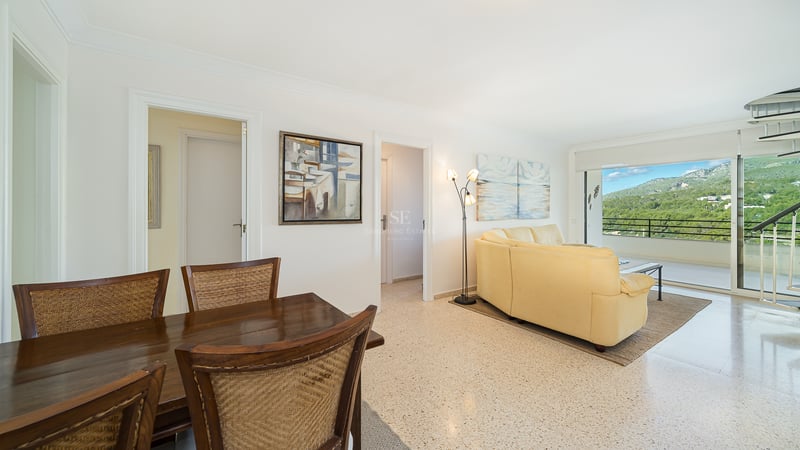 Open living space with a cream leather sofa, wooden dining set, and large glass doors revealing a mountain landscape.