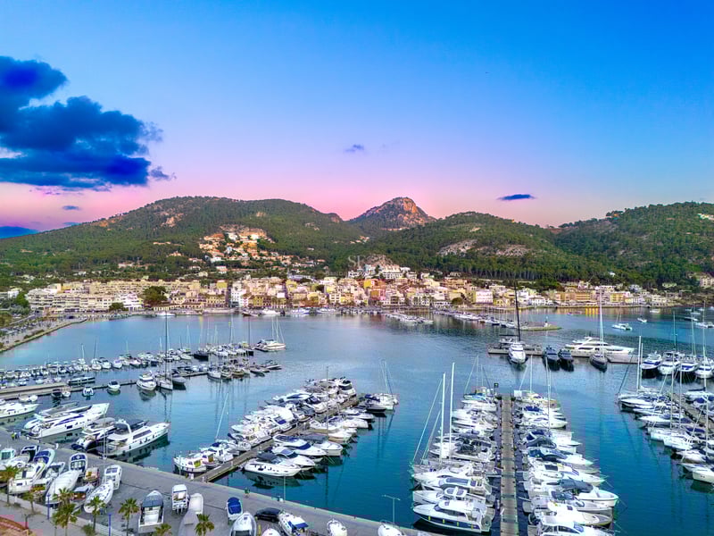 Aerial view of a luxury marina filled with white yachts under a pink and blue sky next to green mountains.