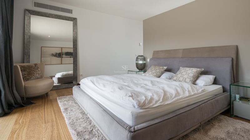 Chambre moderne avec lit gris rembourré, parquet en chêne, grand miroir au sol et tables de chevet en verre.