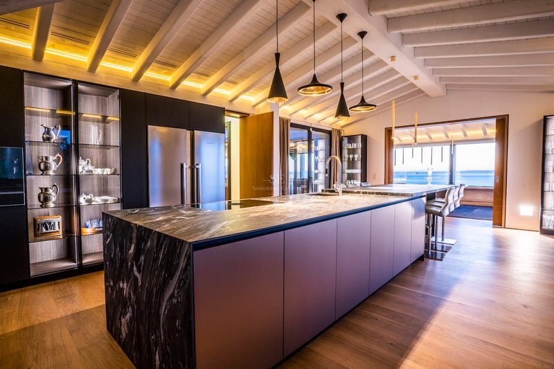 Moderne Küche mit großer Marmorinsel, Holzbalkendecke und Blick auf das Meer durch die Fensterfront.