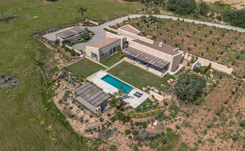 Luftaufnahme einer modernen Steinvilla mit türkisem Pool, grünem Rasen und umgebender Naturlandschaft auf Mallorca.