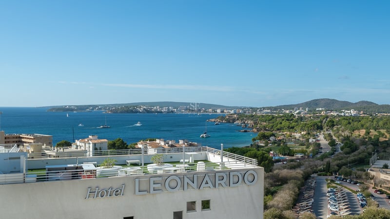 Panoramic view of a Mediterranean bay and coastline from a modern rooftop terrace with white railings and artificial turf.