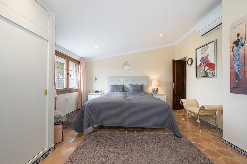 Chambre avec sol en terre cuite, grand lit avec couvre-lit gris, placards intégrés blancs et fenêtre en bois.