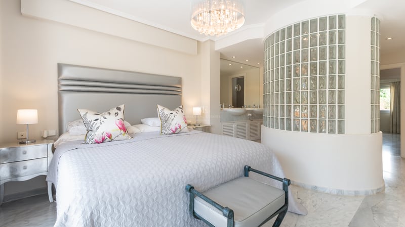 Luxury bedroom with a king bed, marble floors, a crystal chandelier, and a curved glass block wall leading to an en-suite.