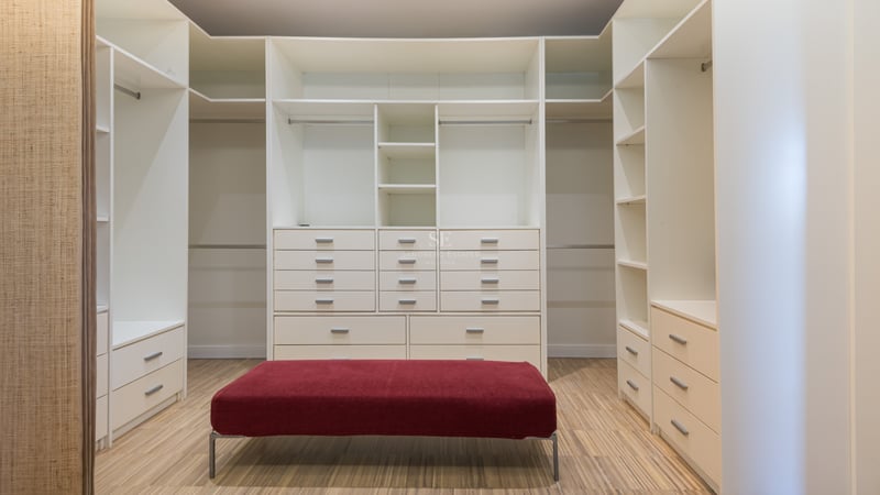 Rymlig vit walk-in-closet med hyllor, lådor, trägolv och en central röd sammetspuff.