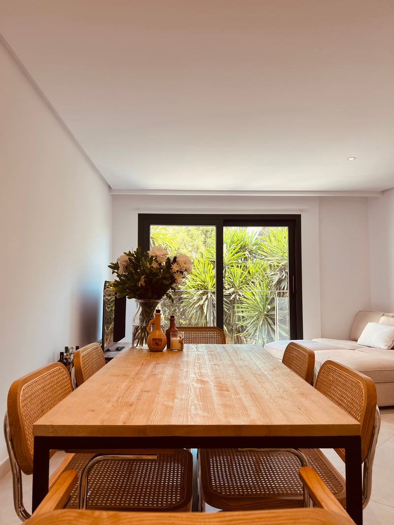 Table à manger en bois clair et chaises en rotin devant une grande baie vitrée donnant sur de la verdure.