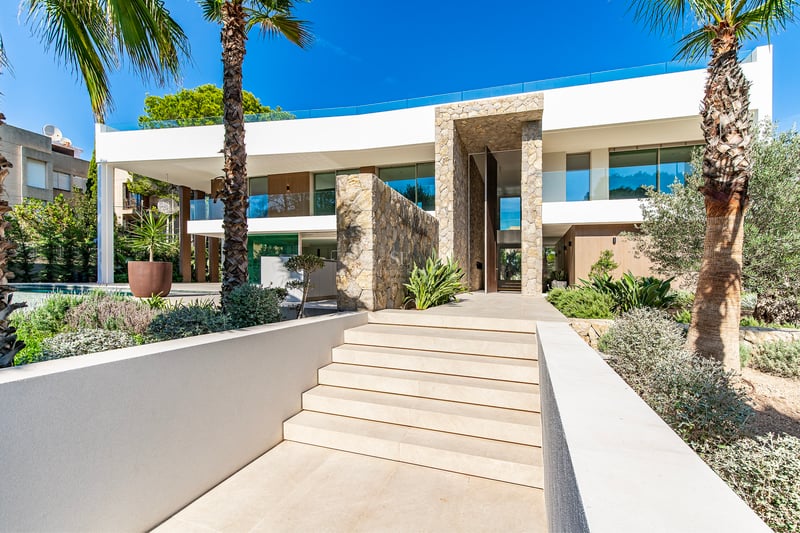 Entrée de villa blanche avec murs en pierre, escaliers extérieurs et palmiers sous un ciel bleu.
