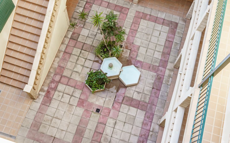 Flygvy över en stenlagd innergård med en hexagonal fontän, växter och en trappa.