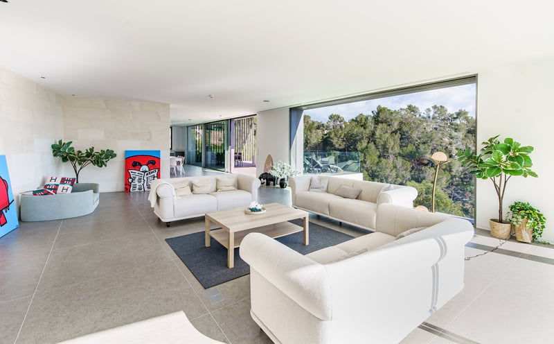 Grand salon moderne avec canapés crème et immenses baies vitrées donnant sur une forêt verdoyante.