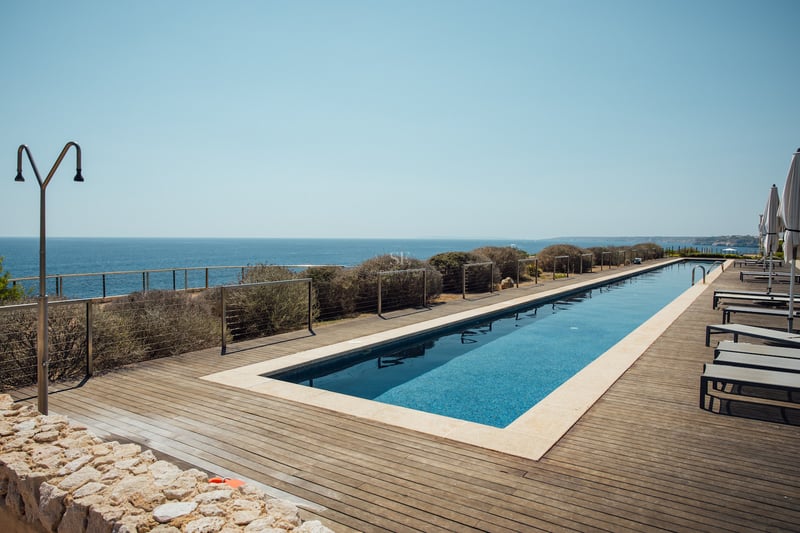Longue piscine rectangulaire sur terrasse en bois surplombant la mer Méditerranée avec chaises longues et douche.