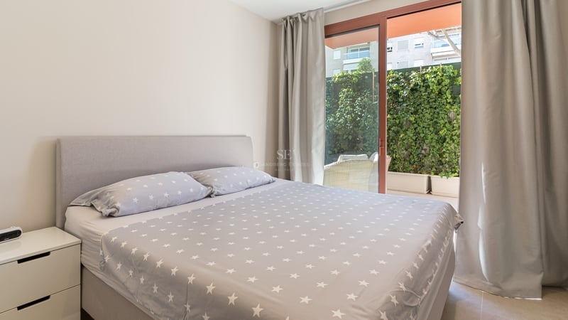 Bright bedroom featuring a grey upholstered bed and sliding glass doors opening to a terrace with lush green hedges.
