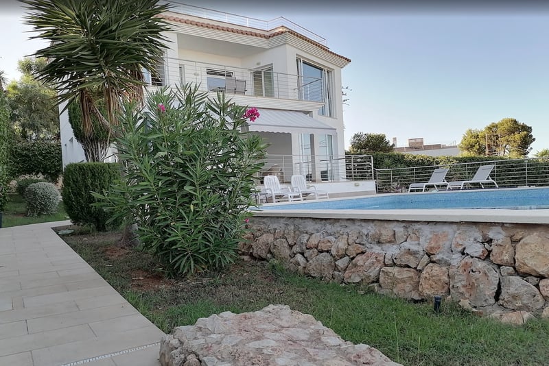 Villa blanche à deux étages avec piscine turquoise, chaises longues et mur de jardin en pierre naturelle.