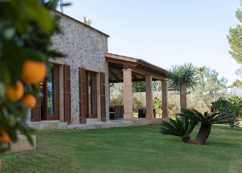 Naturstensfasad med övertäckt terrass, välskött gräsmatta och apelsinträd i dagsljus.