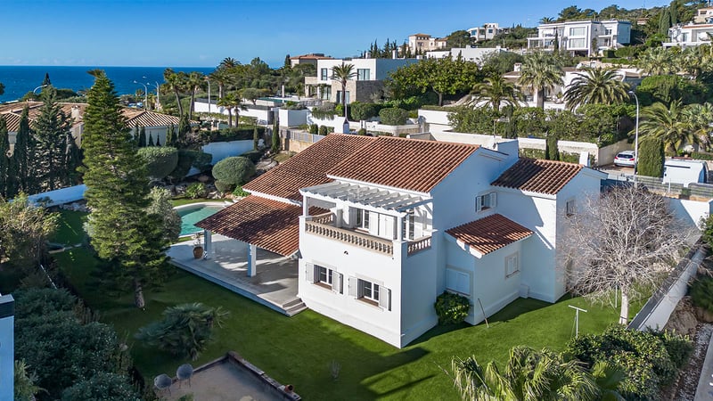 Flygvy över en vit medelhavsvilla med tegeltak, trädgård, pool och havet i bakgrunden under blå himmel.