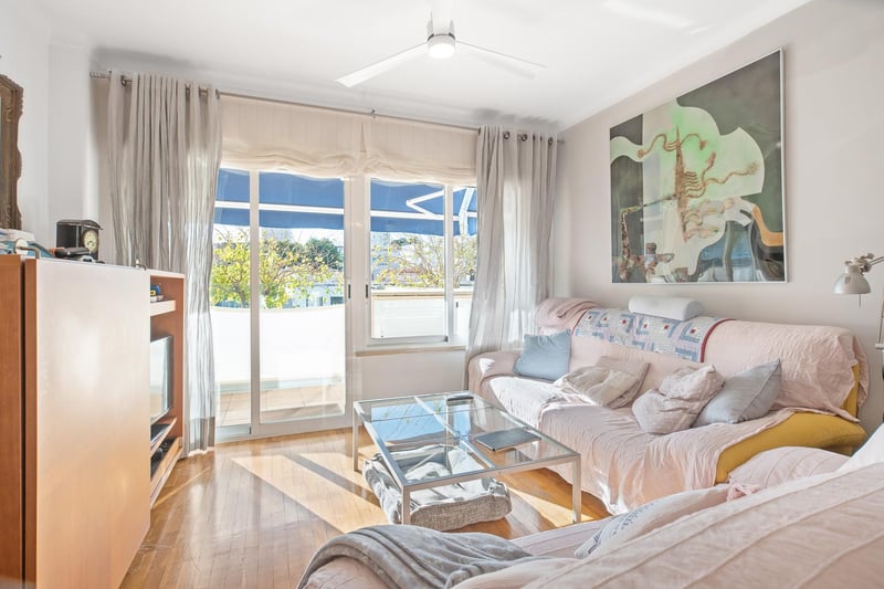 Lichtdurchflutetes Wohnzimmer mit Eichenparkett, Glastisch, rosa Sofa und Glasschiebetüren zum Balkon.