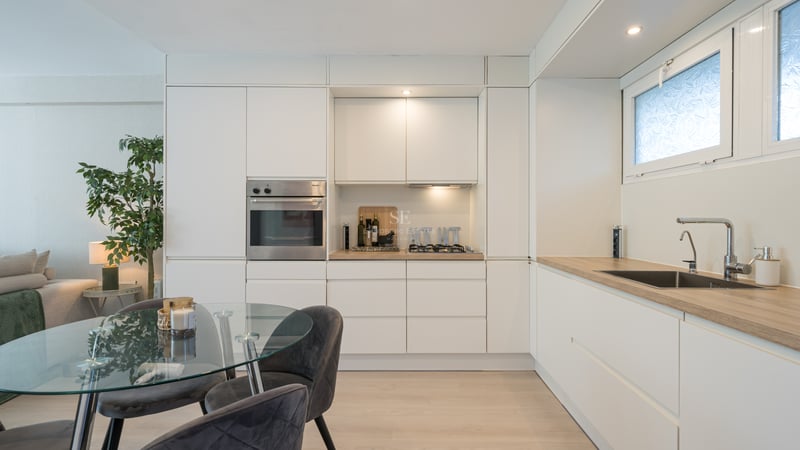 White minimalist kitchen with integrated Miele oven, wood-effect countertops, and a glass dining table with velvet chairs.