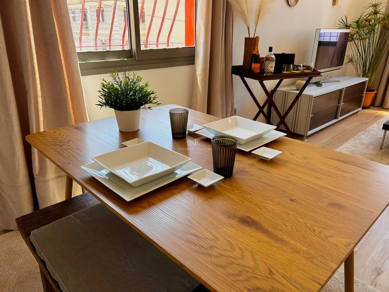 Wooden dining table set with white square plates and glasses in a bright living room.