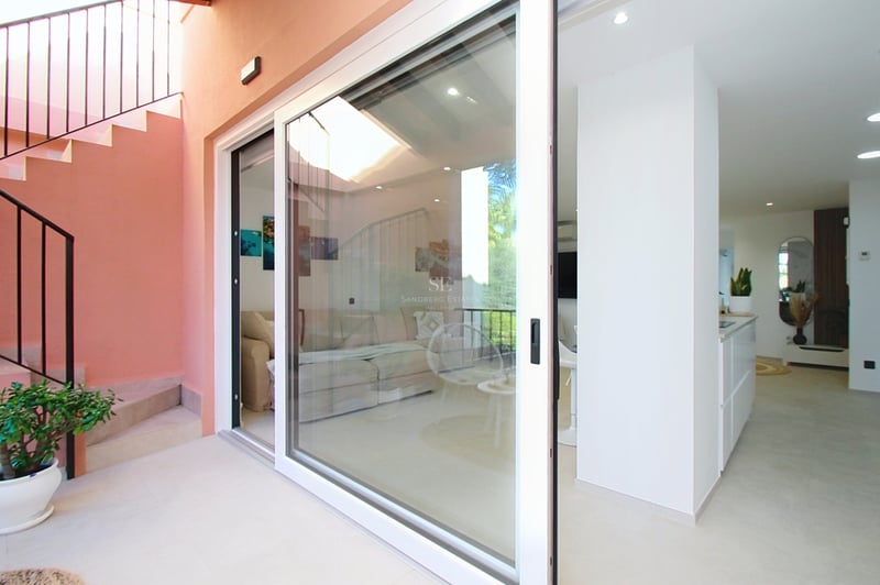 Vy från terrassen genom stora vita skjutdörrar i glas in till ett ljust modernt vardagsrum och kök.