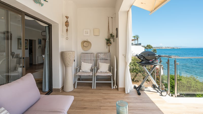 Solig övertäckt balkong med golv i träimitation, sittgrupp, grill och glasräcke med utsikt över det blå havet.