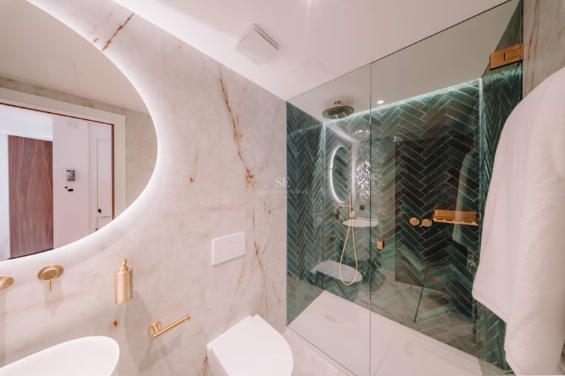 Salle de bain moderne avec murs en marbre, miroir rond rétroéclairé et douche avec carreaux verts en chevrons.