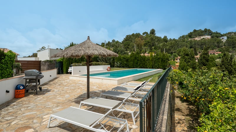 Piscine rectangulaire entourée d'une terrasse en pierre, chaises longues blanches et parasol en paille.