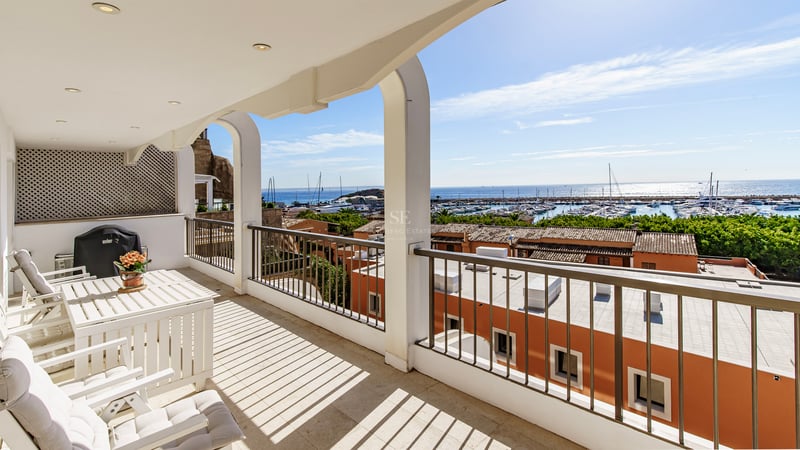 White arched balcony with outdoor furniture overlooking a harbor with boats and blue sea under a clear sky.