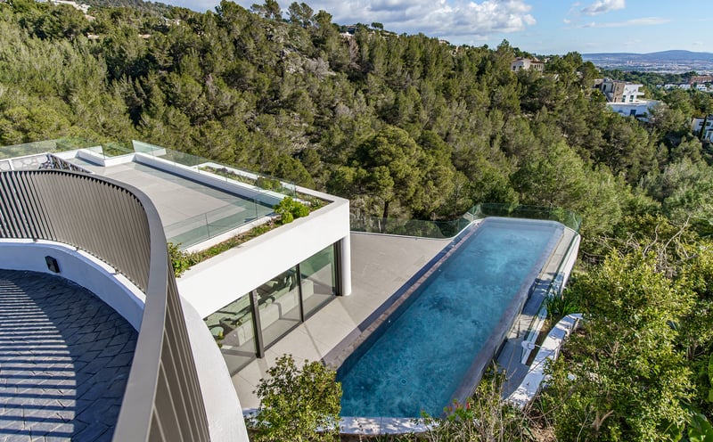 Vue en plongée d'une piscine à débordement d'une villa moderne surplombant une forêt verdoyante sous un ciel dégagé.