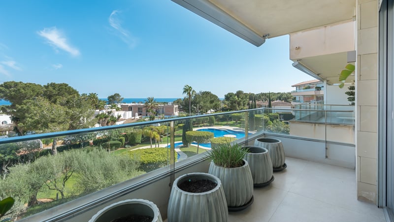 Modern balcony with glass railings overlooking lush green gardens and the blue sea under a clear sky.