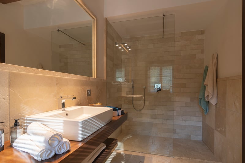 Modernt badrum med duschvägg i glas, kakel i natursten och kommod i mörkt trä med ovanpåliggande tvättställ.