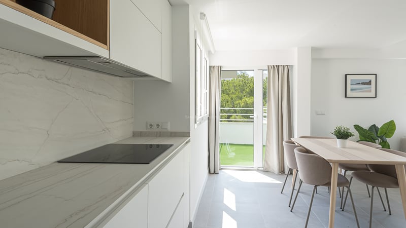 Cuisine blanche moderne avec plan de travail en marbre, salle à manger en bois et porte de balcon.