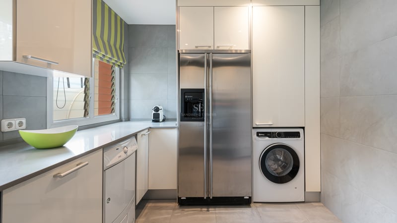 Modern laundry area featuring a large stainless steel refrigerator, washing machine, and white cabinetry against grey tiles.