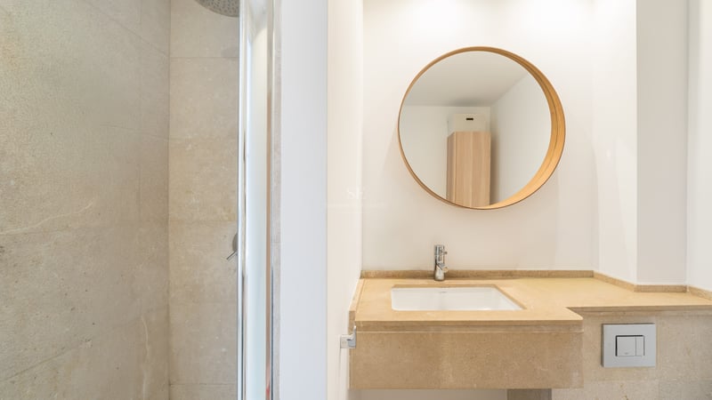 Modernt badrum med tvättställ i beige sten, rund träspegel och duschkabin i glas.