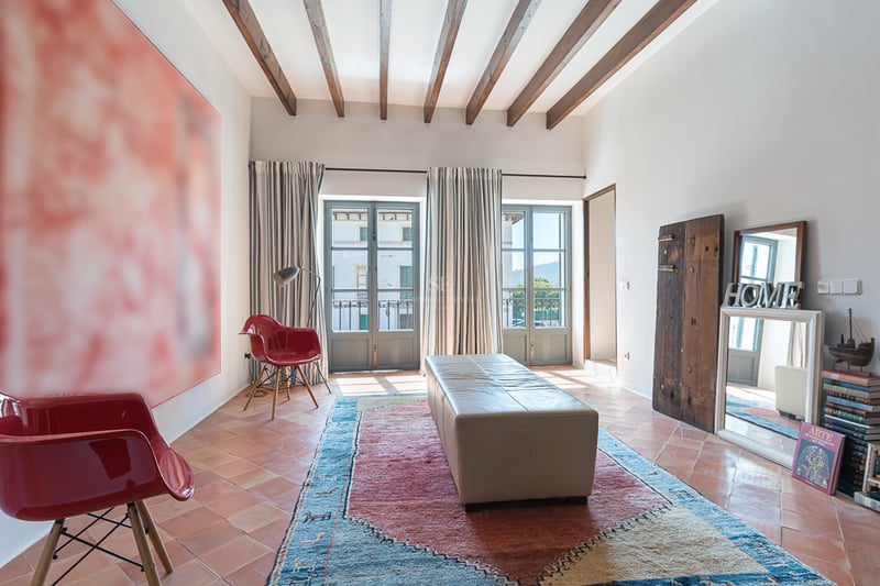 Espace de vie avec sols en terre cuite, poutres en bois sombre, chaises rouges et deux balcons français lumineux.