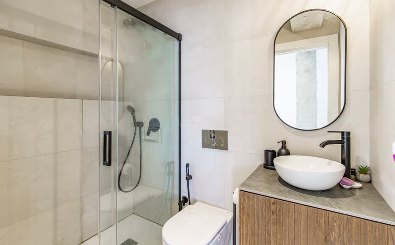 Salle de bain contemporaine avec douche vitrée, robinetterie noire, meuble en bois et miroir ovale.