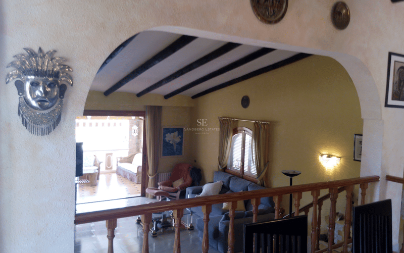 Salón acogedor visto a través de un arco con vigas de madera oscura y acceso a terraza sombreada.