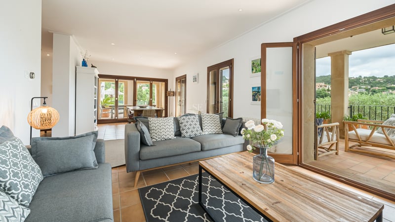 Canapé gris moderne et table basse en bois dans un salon lumineux avec sol en terre cuite et grandes baies vitrées.