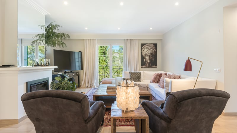 Bright living room with white L-shaped sofa, dark armchairs, wooden coffee table, and balcony overlooking greenery.