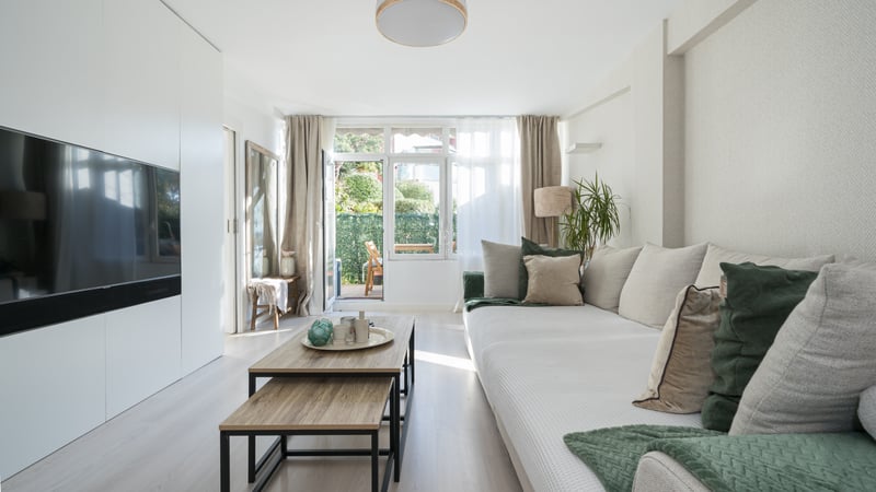 Bright living room featuring a large beige sofa, wooden coffee tables, and glass doors opening to a green terrace.