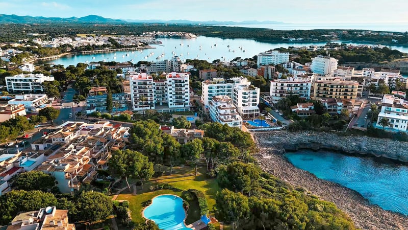 Vue par drone d'une ville côtière méditerranéenne avec des bâtiments blancs, des piscines et une baie bleue avec des bateaux.