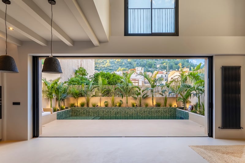 Vue intérieure par une grande baie vitrée vers une piscine privée entourée de plantes tropicales et carrelage vert.