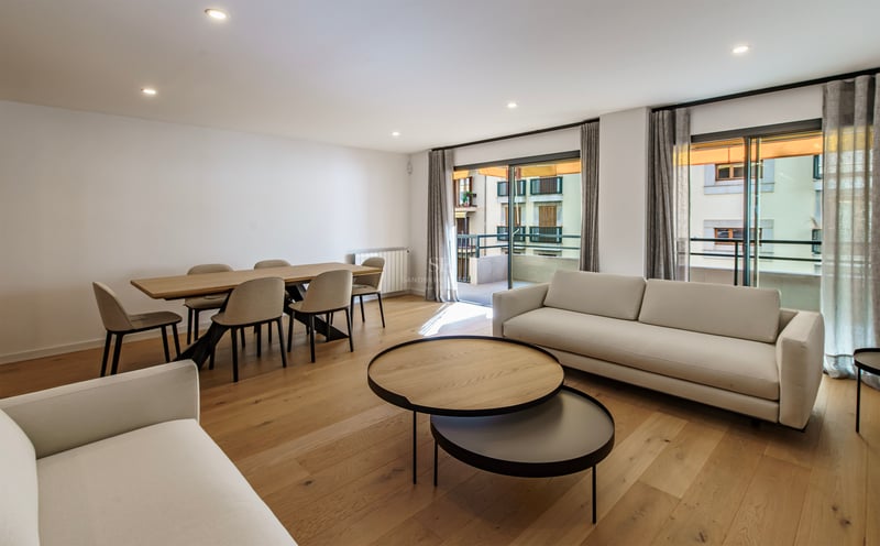 Salon moderne avec canapés blancs, tables basses en bois, coin repas et grandes baies vitrées ouvrant sur un balcon.